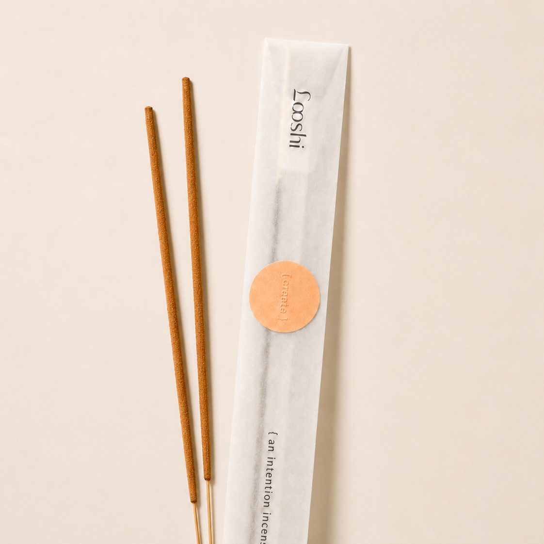 Two incense sticks next to their white paper packaging sleeve with a gold circular sticker, on a light beige background. The sleeve is branded 'Coosii'.