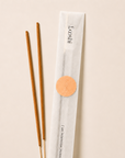 Two incense sticks next to their white paper packaging sleeve with a gold circular sticker, on a light beige background. The sleeve is branded 'Coosii'.