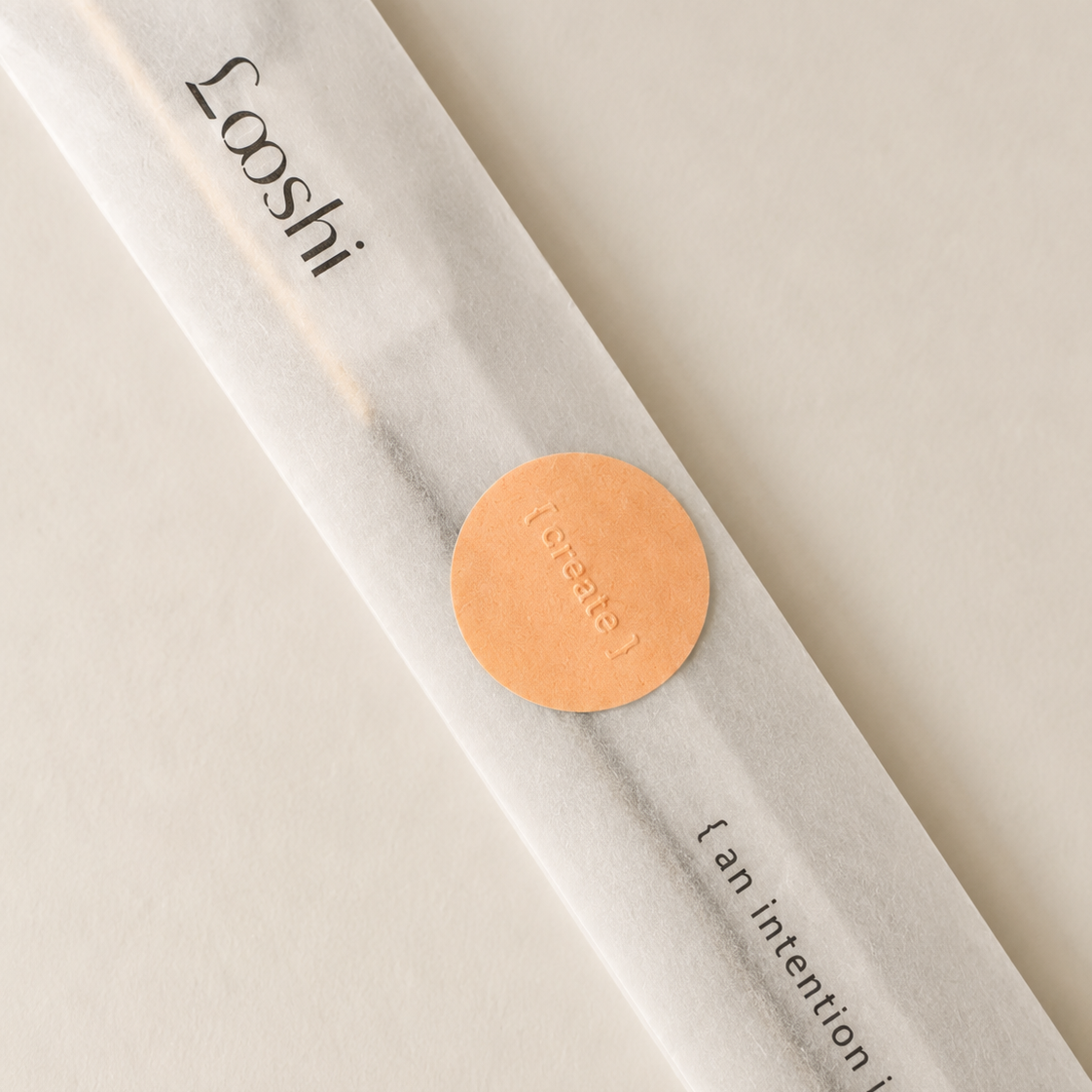 A single incense stick in a translucent white paper sleeve with the brand name 'Looshi' printed in black and a round copper seal, photographed on a warm off-white background.