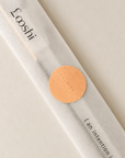 A single incense stick in a translucent white paper sleeve with the brand name 'Looshi' printed in black and a round copper seal, photographed on a warm off-white background.