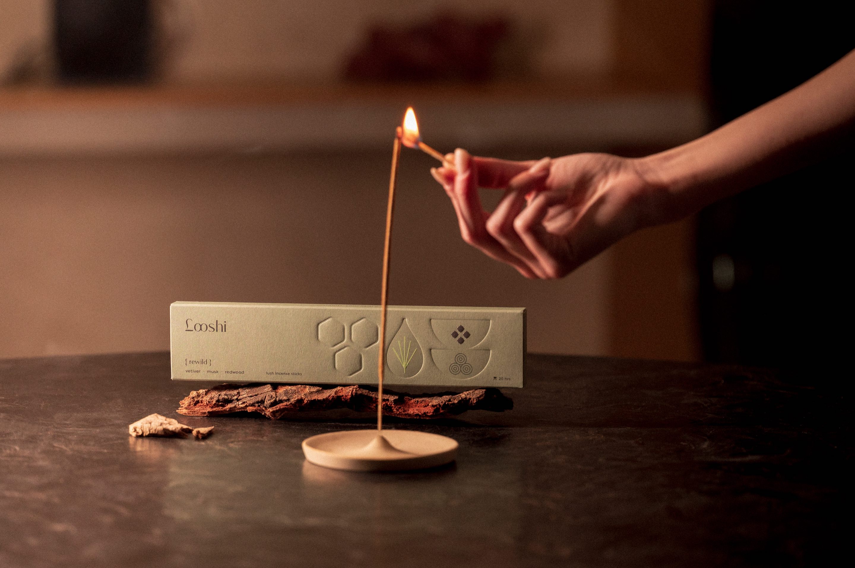 A hand lighting a stick of Rewild beside its box on a piece of redwood bark